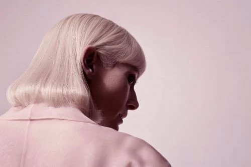 Head and shoulder shot of a woman with platinum blonde hair wearing a pale pink jacket, looking down in a photo studio.