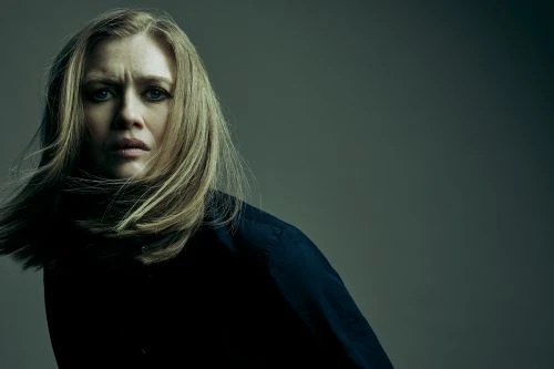Close-up portrait of a blonde woman with a concerned expression wearing a dark top, set against a muted gray background in studio lighting.