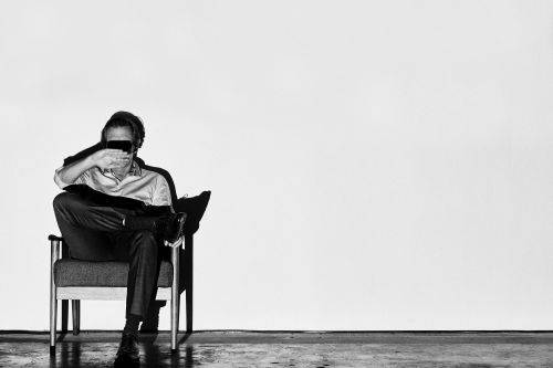 Man seated in an armchair with hand covering his eyes against a plain wall, photographed by James Minchin.