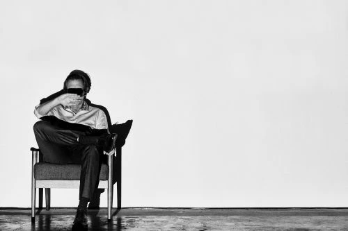 Man seated in an armchair with hand covering his eyes against a plain wall, photographed by James Minchin.