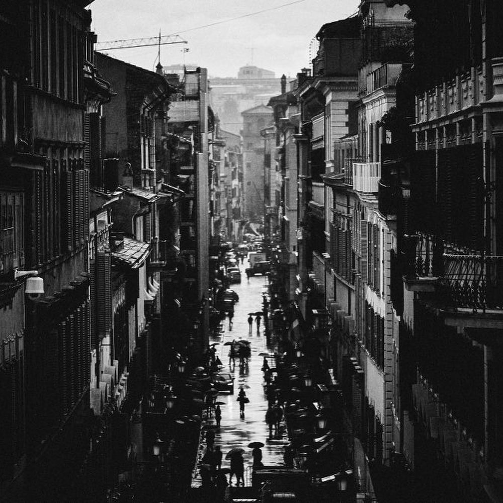 Black and white cityscape photograph of a street in a city with people walking on the wet surface and tall buildings on both sides.