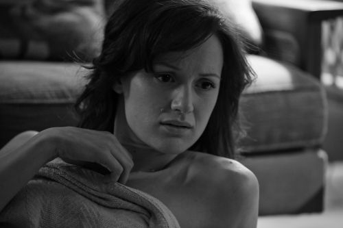 Monochrome portrait of a shoulder-up shot of a pensive adult woman with dark hair, wearing a wrap, sitting indoors with a couch in background.