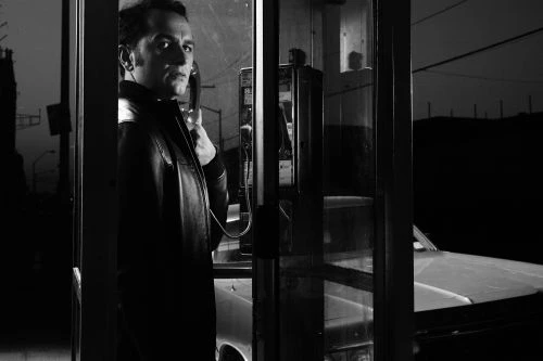 Monochrome portrait of a man in a phone booth wearing a leather jacket, holding the phone receiver, with a car visible outside.