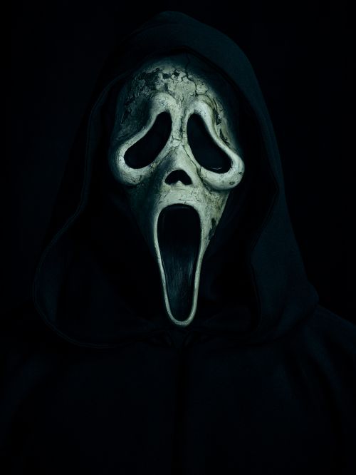 Close-up of a Ghostface mask, the iconic costume from the 'Scream' movies, set against a stark, black background and shrouded in shadow.