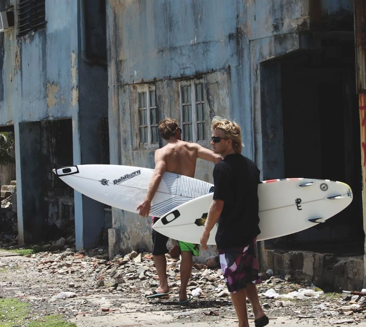 Two shirtless men carry surfboards past derelict building, heading to surf. Outdoor lifestyle, sport, beach. Sunny day, natural light.