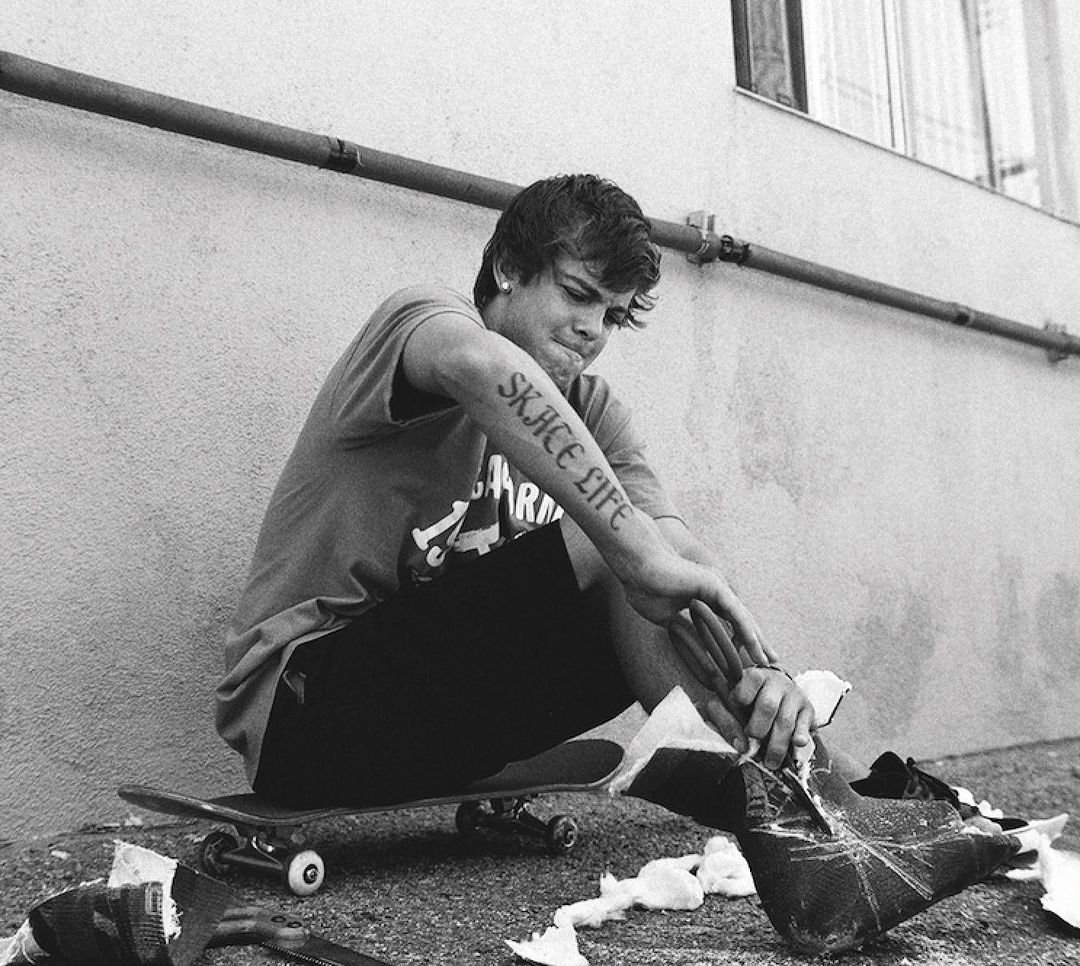 Monochrome shot of a young adult sitting on a skateboard with damaged shoes, featuring a tattoo that reads 'Skate Life'.