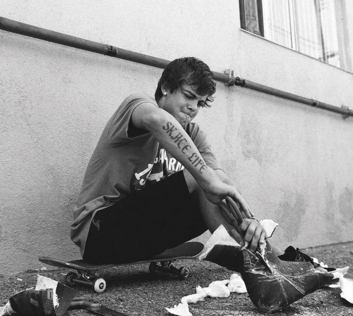 Monochrome shot of a young adult sitting on a skateboard with damaged shoes, featuring a tattoo that reads 'Skate Life'.