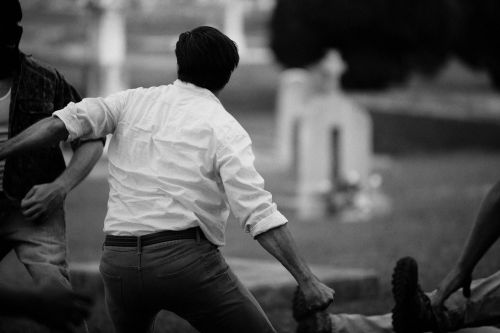 Monochrome image captures 3 adult men in action in an outdoor setting, near tombstones. One is being subdued by the other two.