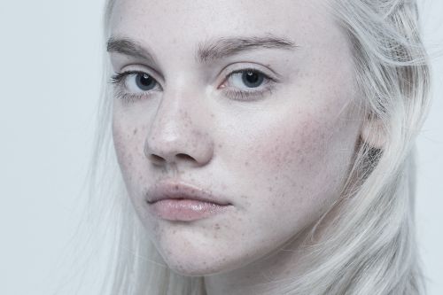 Close-up portrait of a young, fair-skinned woman with light hair and freckles, against a light grey backdrop in soft, diffused lighting.