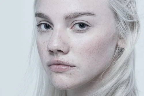 Close-up portrait of a young, fair-skinned woman with light hair and freckles, against a light grey backdrop in soft, diffused lighting.