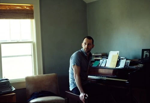 Indoor portrait of a man sitting on piano bench with a typewriter, books, and sheet music visible in a muted, gray-toned room.