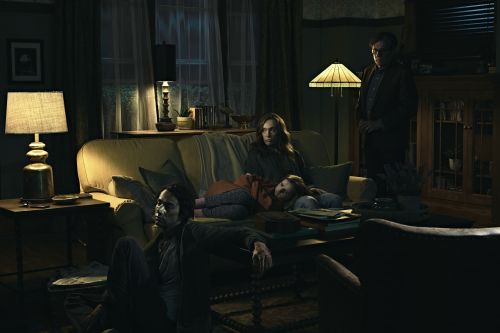 Family in dimly lit living room with child sleeping on couch, man standing near bookcase, mood lighting from lamps.