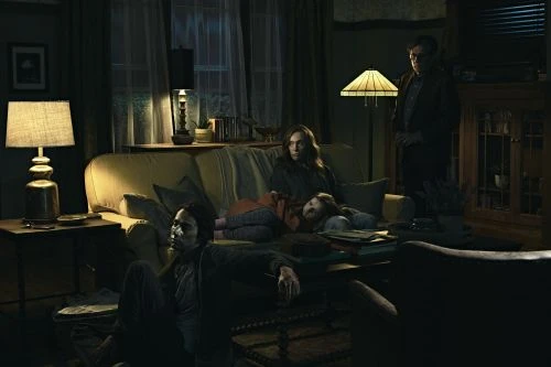 Family in dimly lit living room with child sleeping on couch, man standing near bookcase, mood lighting from lamps.