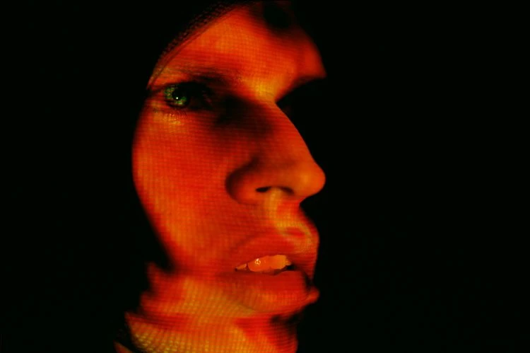 Profile of a person's face with red and orange lighting and a textured overlay, set against a stark black background.