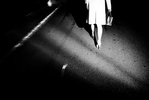 Low angle, black and white shot of woman walking at night wearing a coat and heels, carrying a purse on an empty street.