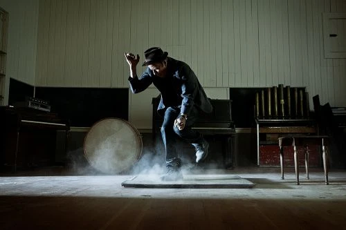 A male tap dancer performs on stage wearing a hat and blazer. The background features musical instruments and smoke on the floor.