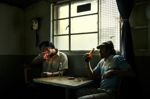 Two men sit at a table by a window, one lighting a cigarette, the other holding a bottle, in a dimly lit, indoor setting.