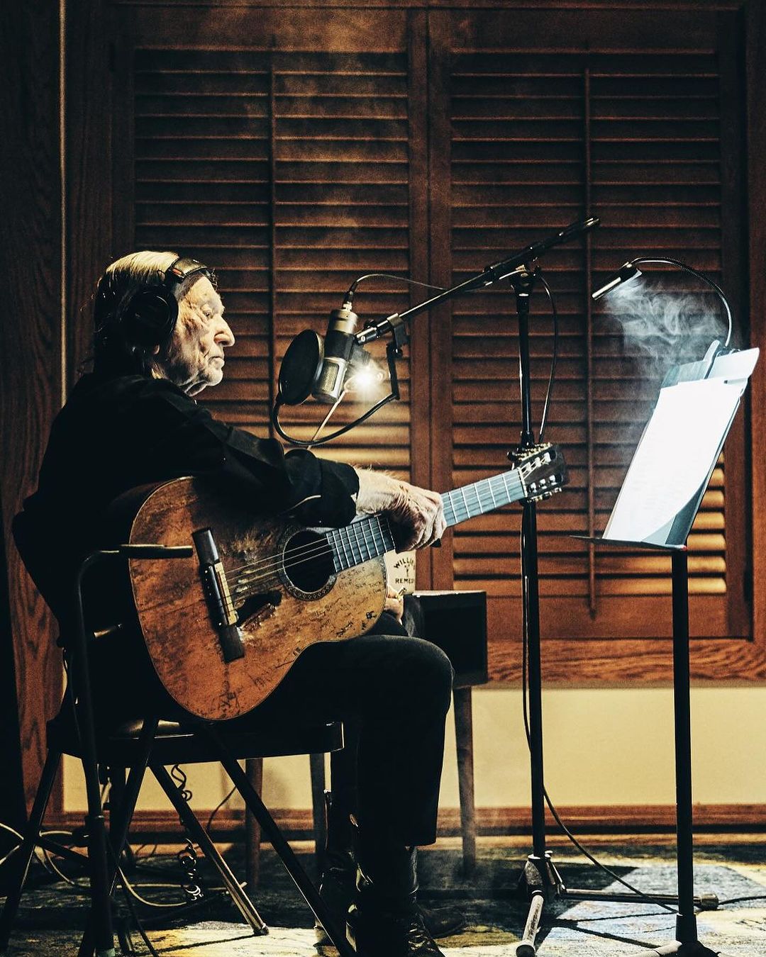 Elderly musician with headphones playing acoustic guitar into microphone in recording studio with louvred shutters backdrop.