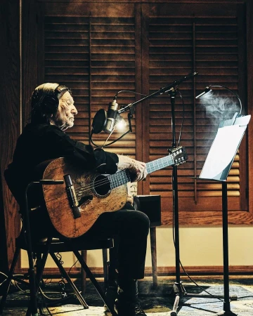 Elderly musician with headphones playing acoustic guitar into microphone in recording studio with louvred shutters backdrop.