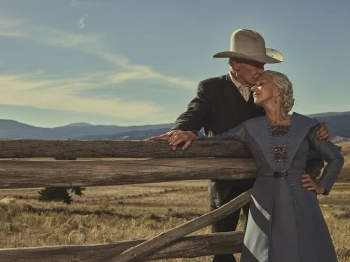 Older couple embraces near a wooden fence against a backdrop of grassy plains and distant mountains under a bright sky.