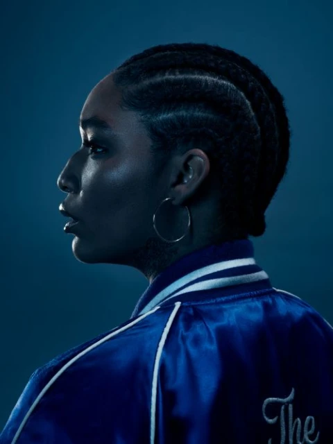 Profile of an adult woman with braided hair wearing a blue satin jacket and hoop earrings against a blue background in a studio.