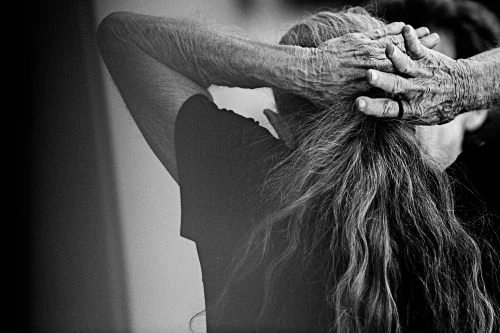 Monochrome portrait of an older woman's wrinkled hands clasping the back of her head, displaying her long graying hair.