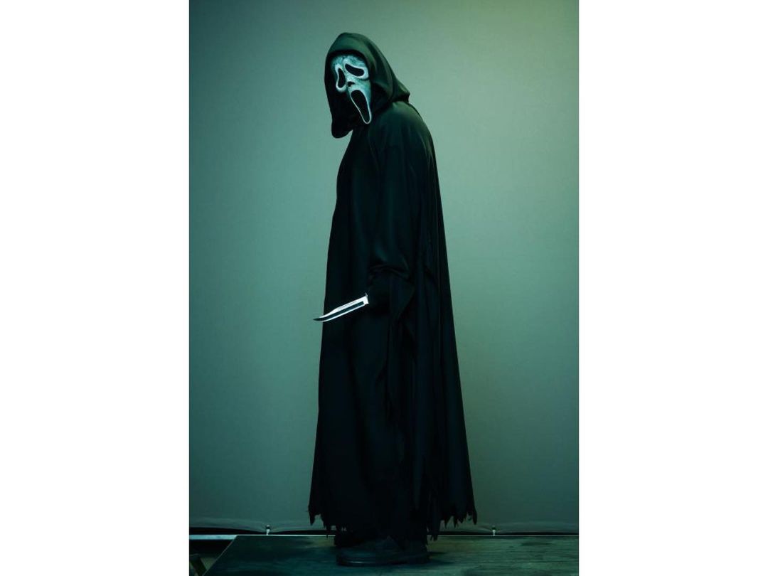 Person in Scream Ghostface mask and black robe, holding a knife, stands in profile against a green backdrop. Horror film concept.