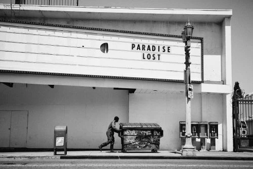 Monochrome image of a man pushing a dumpster past the Paradise Lost theater in an urban setting with a vintage aesthetic.