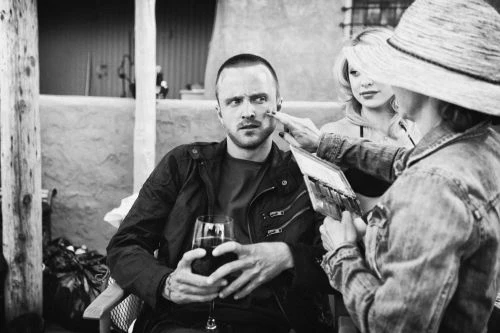 Black and white shot of Aaron Paul as Jesse Pinkman getting makeup applied, with a blonde woman in the background, medium shot.