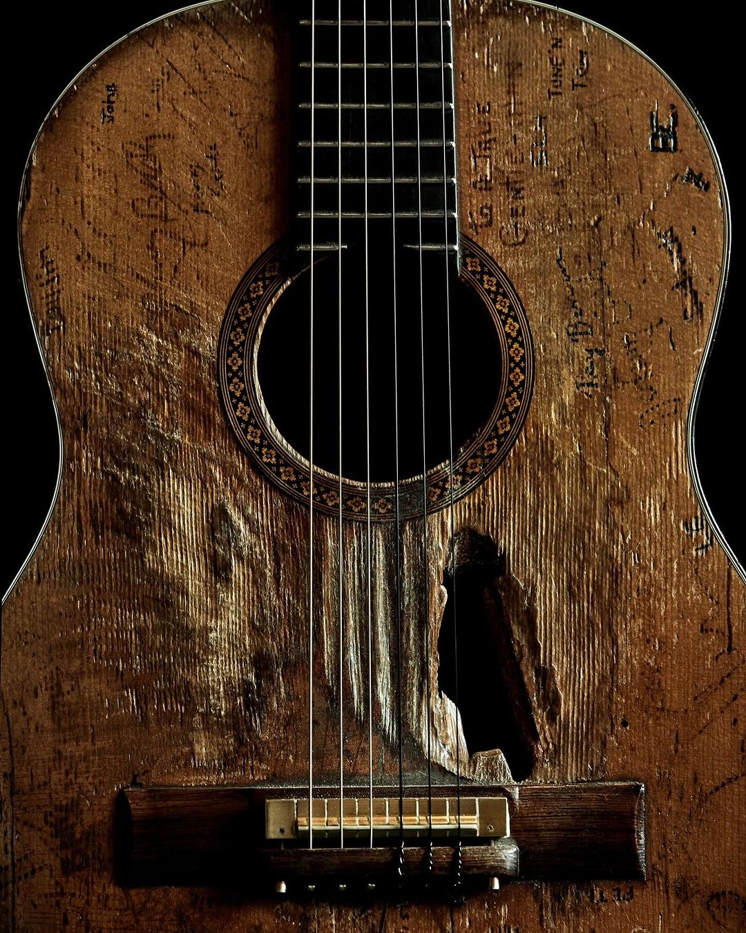 Close-up of an aged acoustic guitar with worn wood texture, a sound hole, and numerous handwritten signatures and markings, set against black.