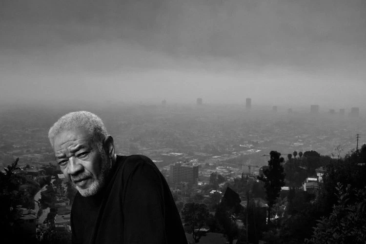 Portrait of an older man in a black shirt with a blurry cityscape backdrop under an overcast sky, in black and white.