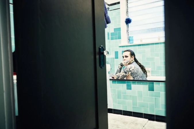 Tattooed adult in bathtub, turquoise tile bathroom, hand to face, serious expression, moody lighting. Doorway framing adds depth.