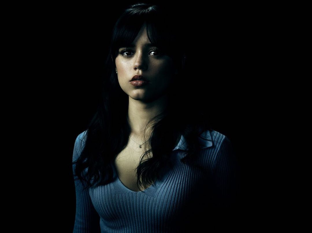 Close-up portrait of a young woman with dark hair and a serious expression, wearing a light blue ribbed shirt against a dark background.