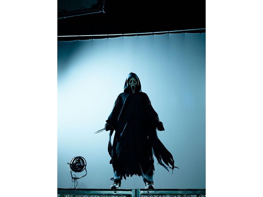 Person in Ghostface costume stands with knife on metal platform against a light blue backdrop, creating a chilling and theatrical image.