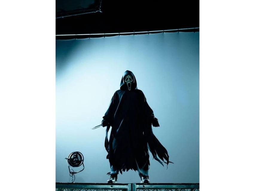 Person in Ghostface costume stands with knife on metal platform against a light blue backdrop, creating a chilling and theatrical image.