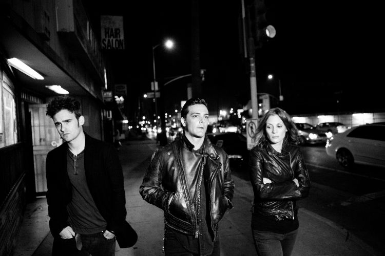 Three people stand on a sidewalk at night in a monochrome image. Two men and a woman in leather jackets are on the city street.