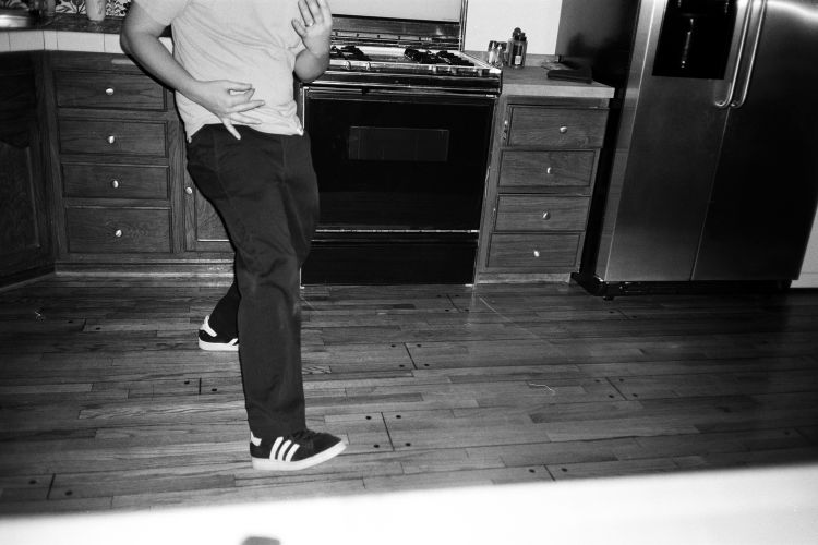 Man in kitchen wearing sneakers and dress pants, hands up, near oven and stainless steel refrigerator. Black and white photography.