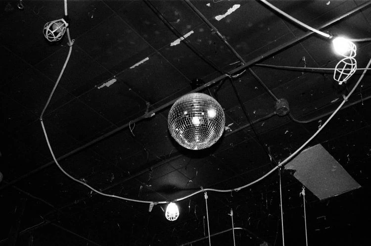 Black and white photo of a mirrored disco ball hanging from a ceiling with lights, wires, and pipes, creating a retro nightclub atmosphere.