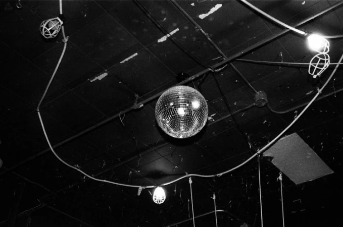 Black and white photo of a mirrored disco ball hanging from a ceiling with lights, wires, and pipes, creating a retro nightclub atmosphere.