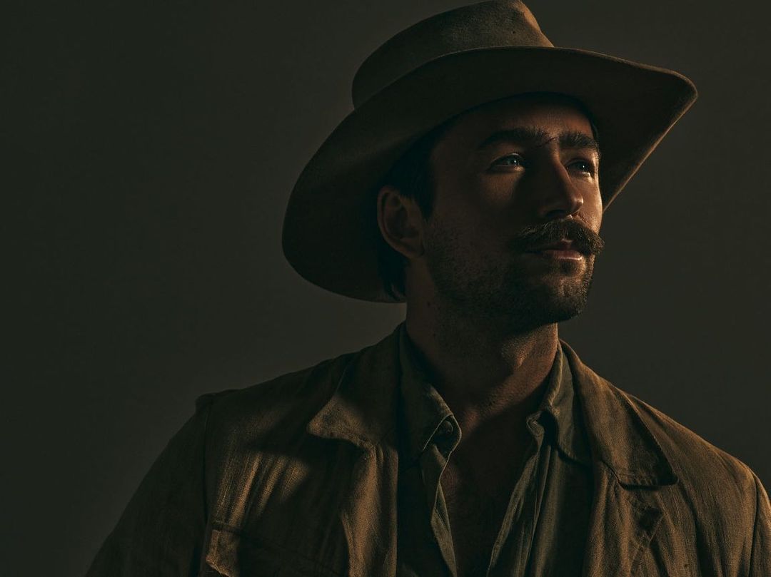 Portrait of adult man with mustache and hat in tan coat, posed against a dark background in muted warm light.