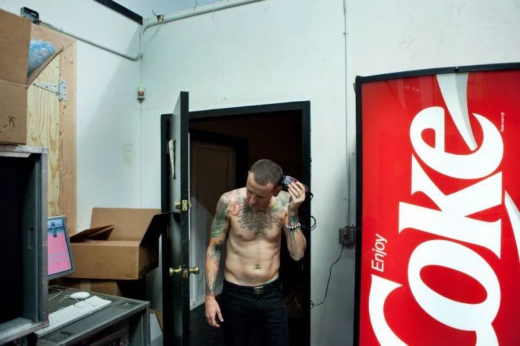 Tattooed man using hair clippers inside a room with a Coca-Cola vending machine, computer and exposed wooden wall panels. 