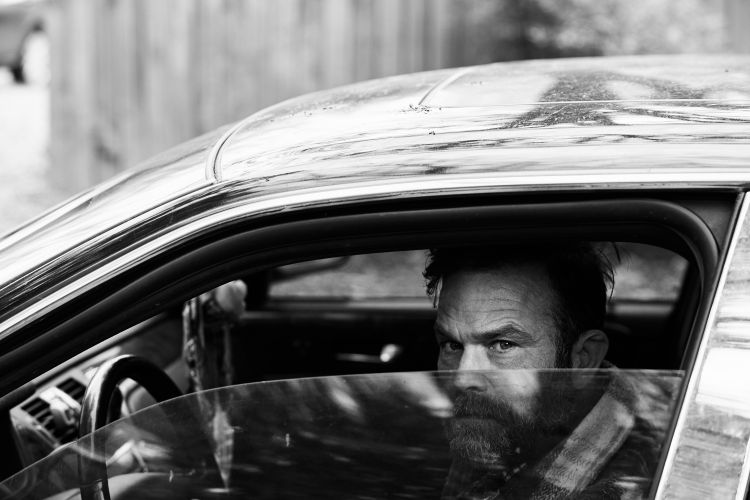 Black and white shot of a bearded man with furrowed brows looking out from inside a car, with a glimpse of the steering wheel.