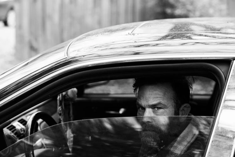 Black and white shot of a bearded man with furrowed brows looking out from inside a car, with a glimpse of the steering wheel.