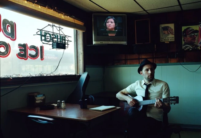 A man in a hat plays guitar in a dimly lit diner setting, with vintage decor, posters, and a TV showing a news program in the background.