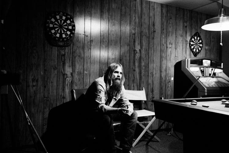 Black and white portrait of a long-haired, bearded man sitting in a game room with a pool table, jukebox, and dartboards.