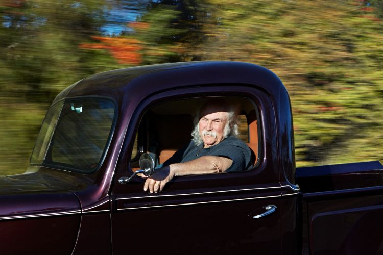 Adult male with long white hair drives a classic pickup truck on a rural road with green trees and motion blur in the background.
