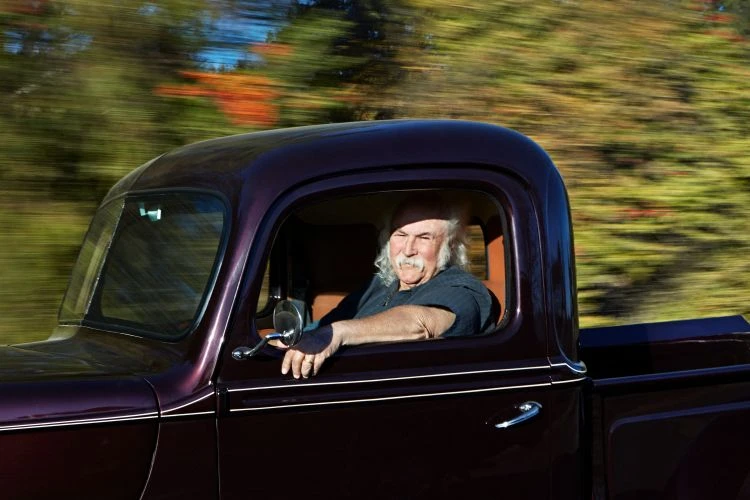 Adult male with long white hair drives a classic pickup truck on a rural road with green trees and motion blur in the background.