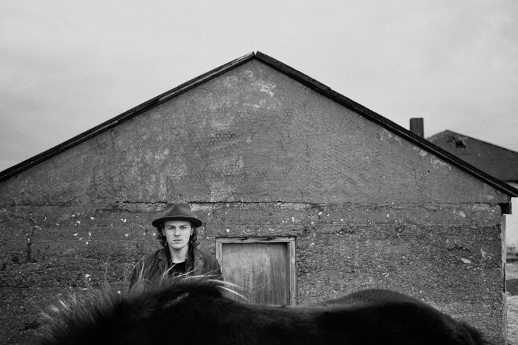 Monochrome portrait of a man in a hat standing outdoors, with a horse in the foreground and a weathered building in the background.