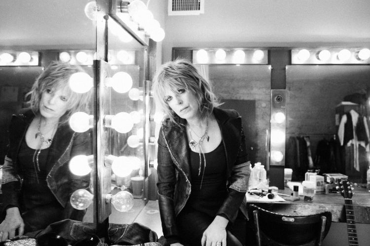 Monochrome portrait of a blonde woman in a black jacket sitting at a vanity mirror with light bulbs, exuding a classic rock vibe.