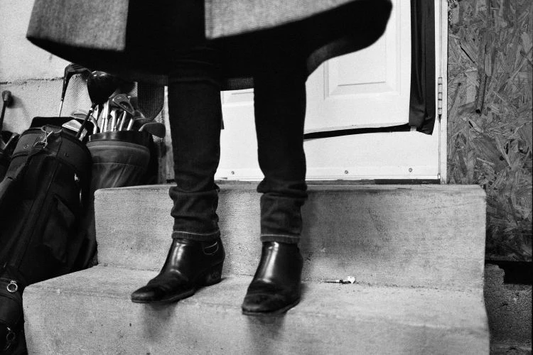 Person in black pants and boots standing on steps next to golf bag, clubs, and a partially opened white door in a black and white image.